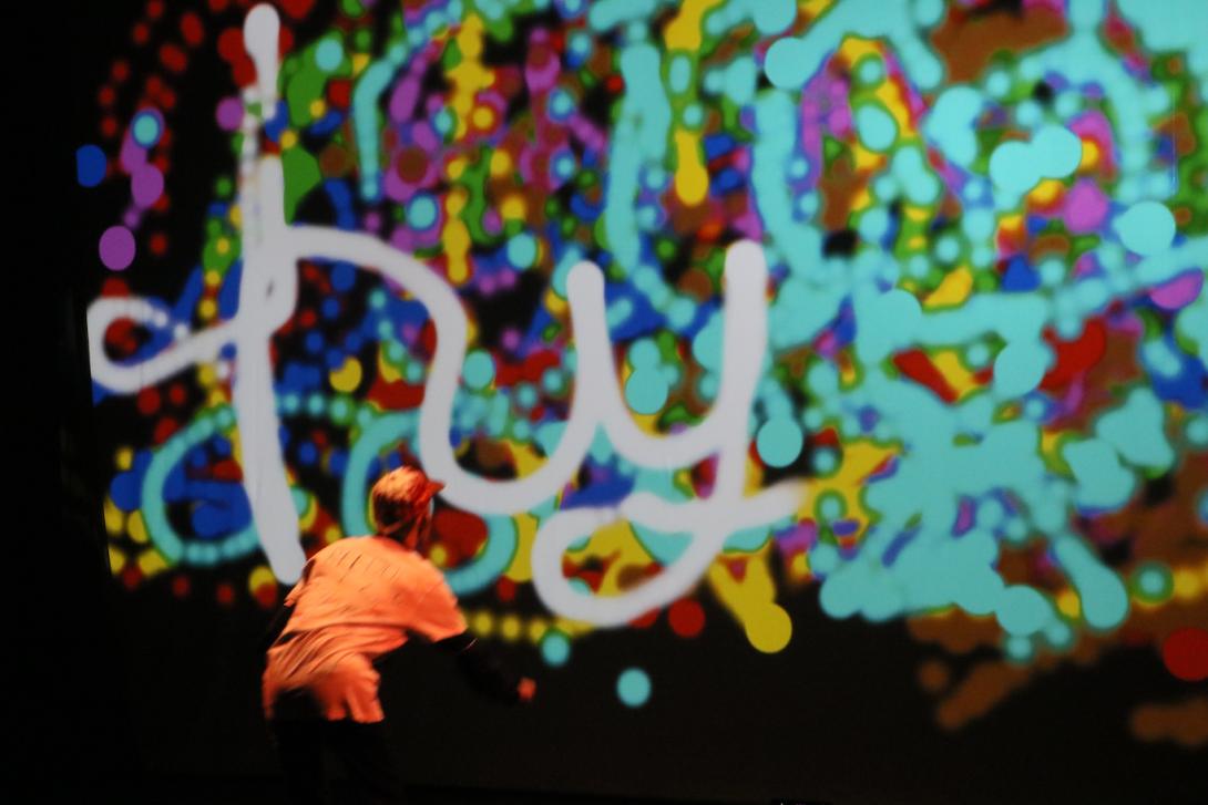 Photo taken during a performance of Hyper. A dancer, bent forward at the waist, faces away from the camera and toward a projection screen, on which he's painting the word 'hyper' in cursive.