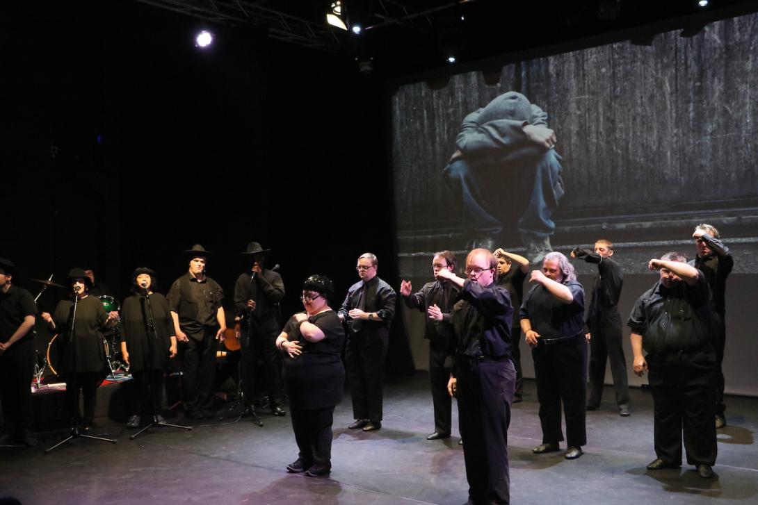 Photo taken during a performance of Hyper. Nine dancers stand in formation making an arm gesture. Several signers with microphones stand in a row off to the side. All are dressed in black. On a projection screen behind the performers is a photo or video of a young person in hoodie and jeans sitting with their head down.