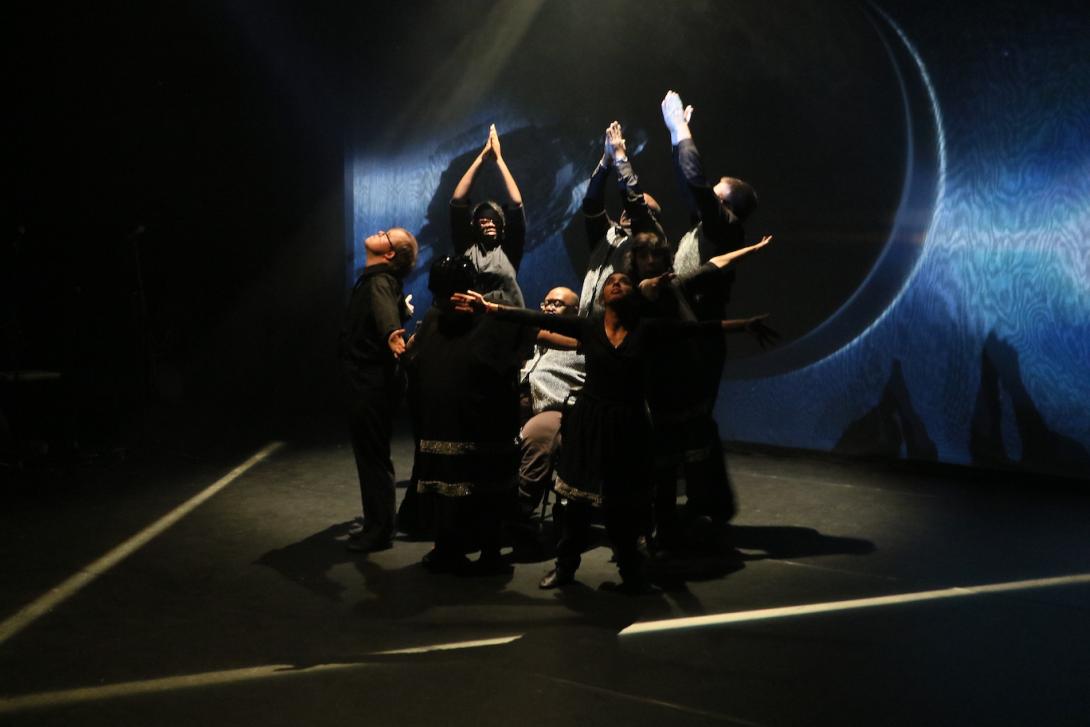 Photo taken during a performance of Hyper. A group of several dancers pose: some with arms steepled above head, some with arms outstretched to the sides, and one kneeling.