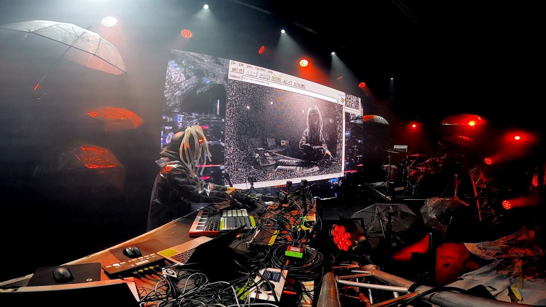 A musician performing onstage with a multitude of electronic equipment, wearing a helmet with clear tubes projecting like dreadlocks, in front of a screen on which a stylized camera feed of the musician is shown inside a Windows 98-style window