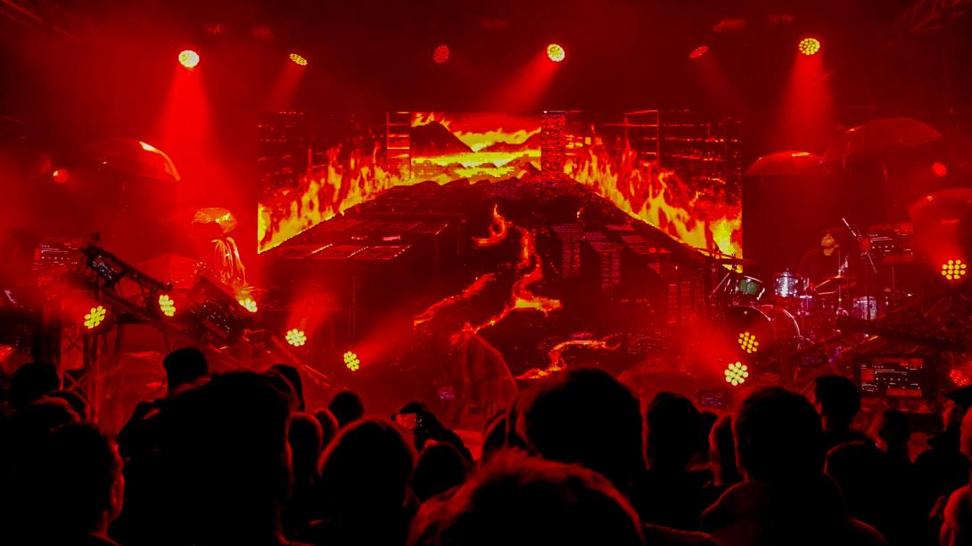 Musicians performing on a stage with a fiery backdrop displayed on a screen; red lights pointing in different directions, equipment, and umbrellas on stage; as seen from behind the heads of a few dozen audience members