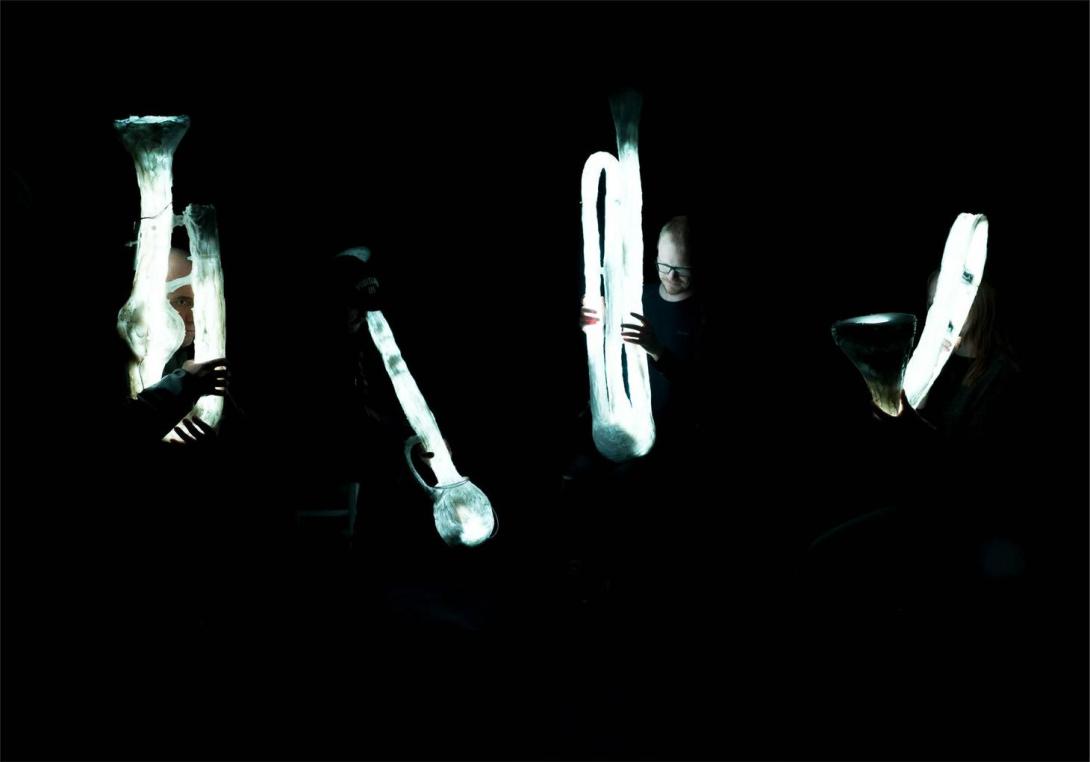 Still from 'Trust me tomorrow' showing four musicians in the dark holding large brass instruments that give off a soft white glow.
