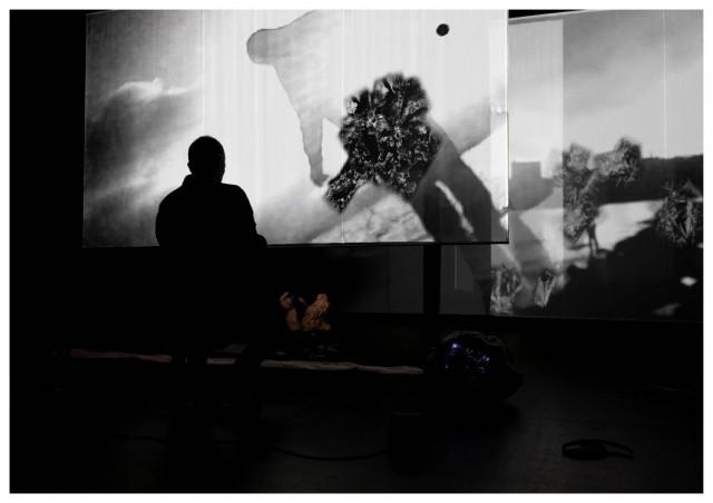 Still from "Trust me tomorrow" showing a silhouette of a human in front of two projected black-and-white images of abstract, overlapping forms.