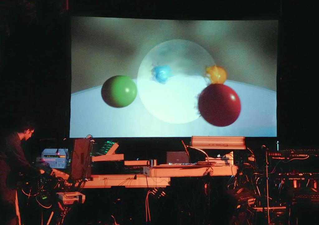 A photo of an electronic music performance with 3D projected visuals of several colorful spheres