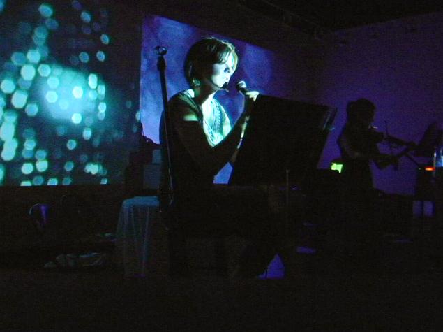 A photos of performers in an art gallery, speaking and playing an electric viola in front of projected video imagery