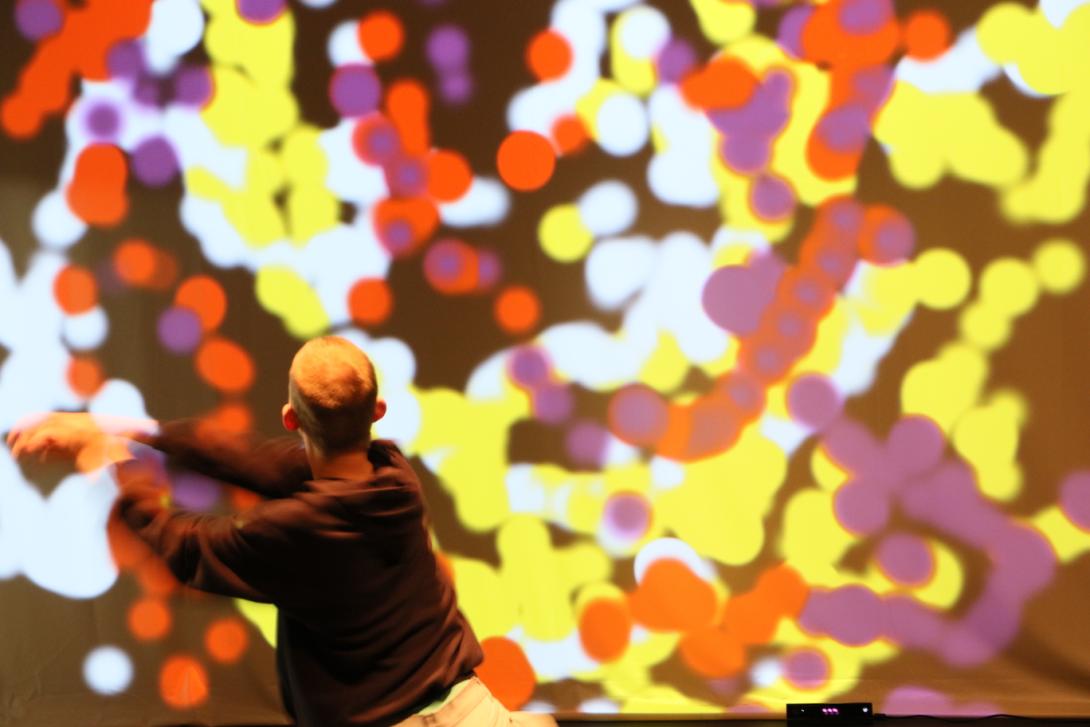 Photo taken during a performance of Hyper. A dancer with his back to the camera extends his arms to the left. In front of him is a projection screen with dots of various colors overlapping and forming trails.