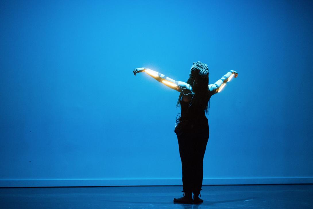 Photo of Kendra Ross wearing LED strips on her outstretched arms and facing a wall bathed in blue light