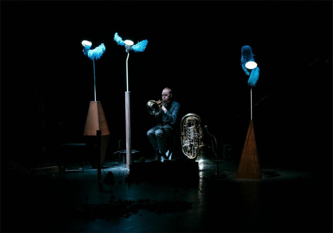 Still from 'Trust me tomorrow' showing a horn player seated on stage  with an extra large brass instrument placed beside him and three tall lamps surrounding him, each with two blue feathers.