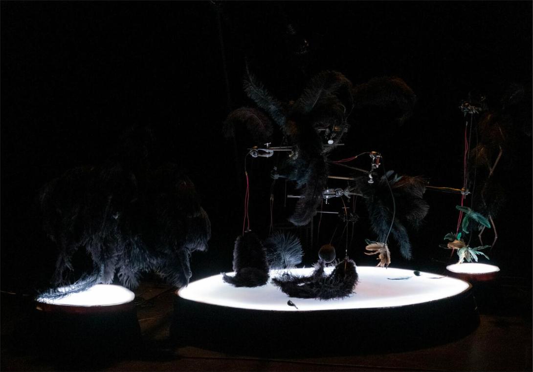 Still from 'Trust me tomorrow' showing a close up of a section of the stage with three lighted circular platforms of different sizes holding a number of mechanical contraptions that all end in feathers.