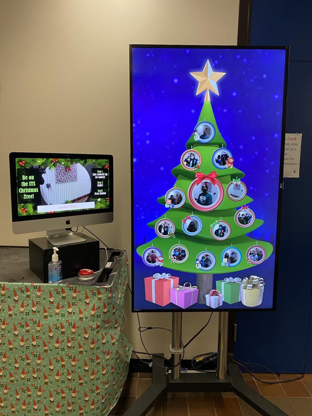 A tall monitor displays a cartoon Christmas tree decorated with circular ornaments containing photos of people. Next to it, a smaller monitor rests on an AV cart decorated with Christmas wrapping paper. The monitor displays instructions for taking a photo. Next to the monitor are a large red button labeled 'easy' and a bottle of hand sanitizer.