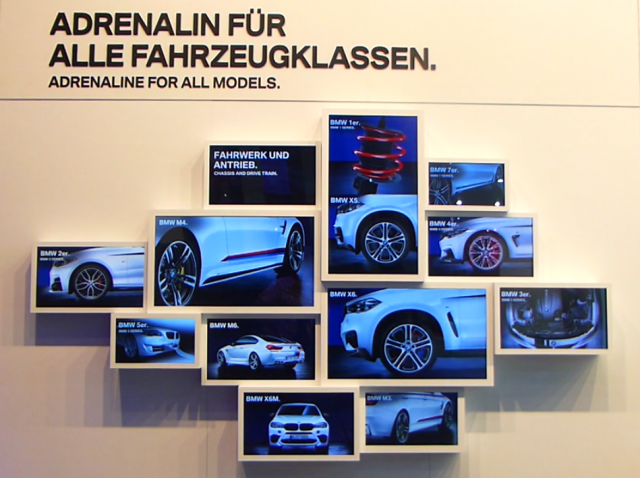 A photo of the wall of a trade show booth, with many monitors showing images of a car