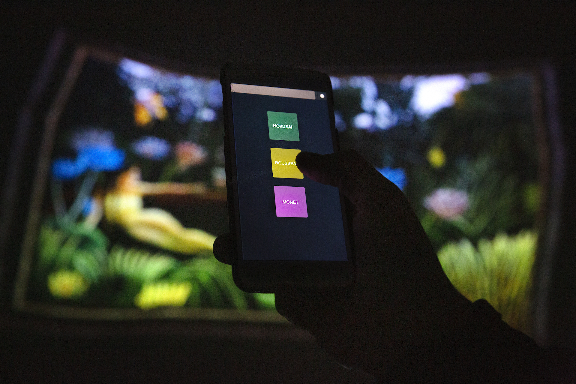 A hand holding a phone showing the app that runs the fulldome projections, with three buttons labelled Hokusai, Rousseau, and Monet. An unfocused image of a fulldome projection of Rousseau's The Dream is the backdrop.