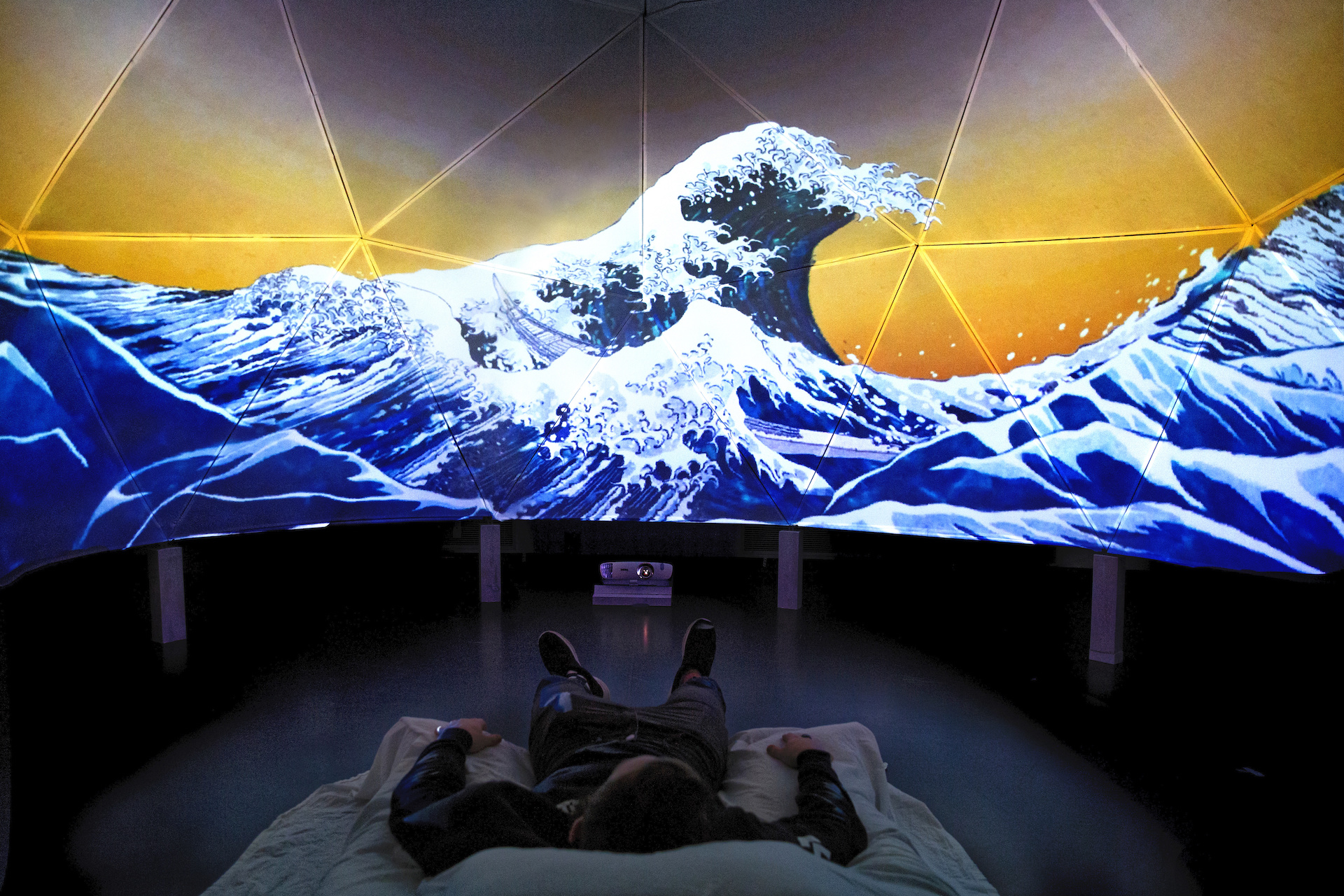 Hokusai's The Great Wave off Kanagawa presented on a large dome screen made of connected triangles. Below, a figure lies on a bed looking up at the projection.