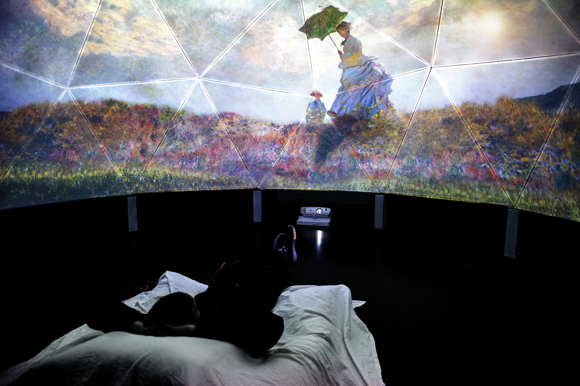 Art by Monet projected on a large wall.  On the floor are a projector and a visitor lying on a bed, looking up at the projection.