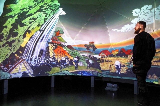 A bearded person stands inside of a dome (5m diameter, 3m tall) looking at the projection on the dome's interior of a composite image of Hokusai prints. Along the floor are a projector and the legs supporting the dome.