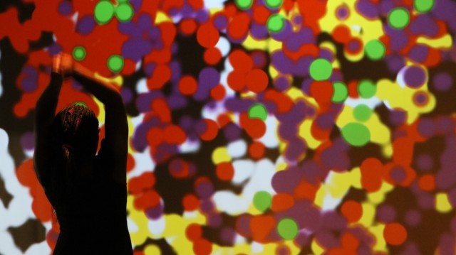 A dancer with arms above head, silhouetted against a spattering of large multi-colored dots on a projection screen