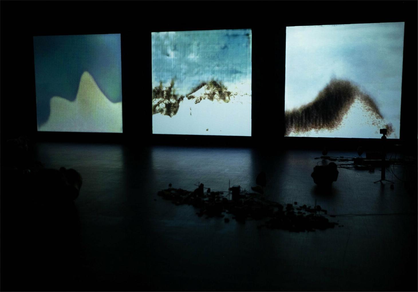 Still from 'Trust me tomorrow' showing an unlit stage with machinery just visible in the foreground but focus drawn to three lit art panels lining the back wall. The images are primarily shades of blue, white, and black in abstracted shapes that suggest motion.